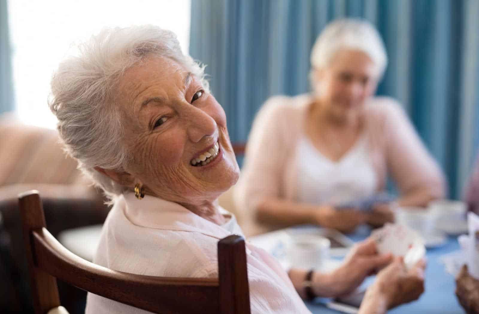 A senior living community resident looking over her shoulder and smiling while playing a game of cards with her peers.